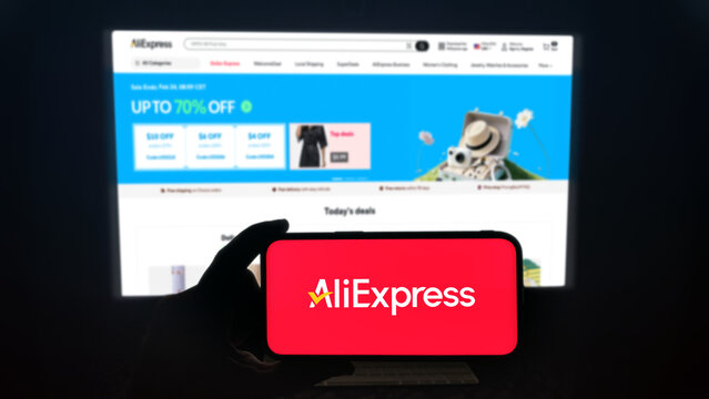AliExpress app running on a modern mobile phone. The smartphone is being held in landscape mode, in the darkness. A PC monitor with the AliExpress website in the background.