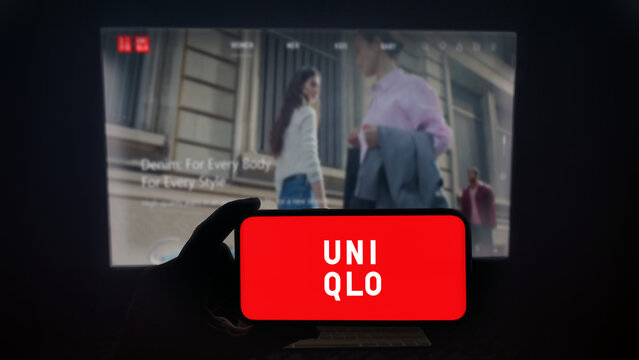 Uniqlo app running on a modern mobile phone. The smartphone is being held in landscape mode, in the darkness. A PC monitor with the Uniqlo website in the background.