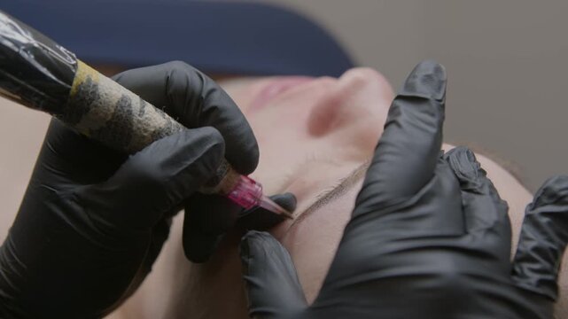 Close-up beauty treatment: Woman undergoes microblading procedure for permanent eyebrow tattoo in salon. Master uses needle tattoo machine for precise shape and natural effect, relaxing atmosphere.