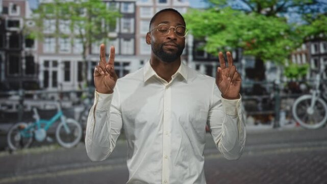 Man makes air quotes with both hands on an amsterdam street by a canal, wearing glasses and a white shirt; sarcasm sly commentary.