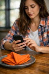 Young woman sitting at wooden table using smartphone with orange napkin on plate in front of her  casual lifestyle JPG image