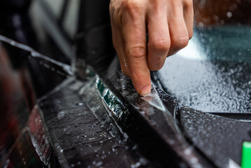 Fototapeta premium A technician peels clear protective film along a trim line on glossy black bodywork, with water beads and moody shallow depth lighting emphasizing precise craftsmanship.