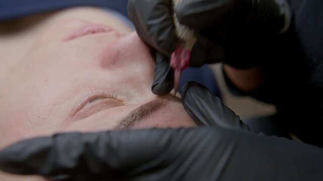 Close-up of microblading procedure: beautician performs permanent eyebrow tattoo using needle machine on woman in beauty salon. Fine strokes for perfect shape and natural look, professional beauty 