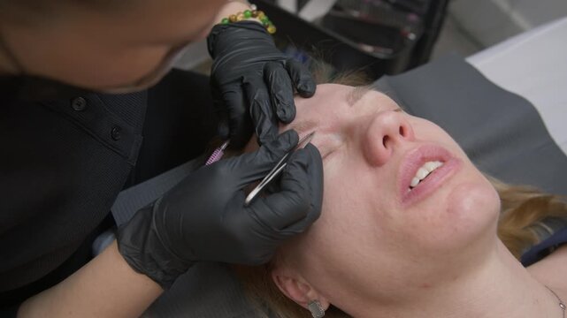 Beautician performs microblading permanent eyebrow tattoo using needle machine on young woman. Natural-looking hair strokes, precise shape correction in professional beauty salon environment.