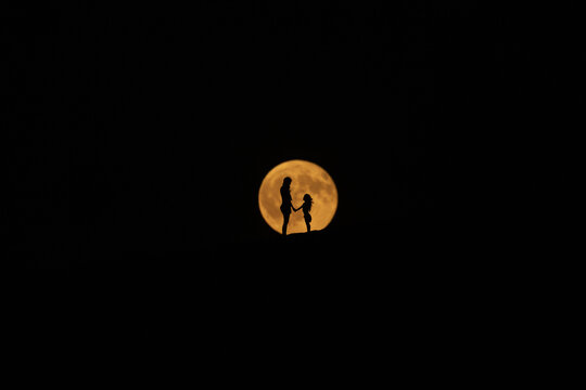 View of a mother and child standing in silhouette against the warm, glowing orb of the moon, capturing an intimate moment in Dune du Pilat, Nouvelle-Aquitaine, France.
