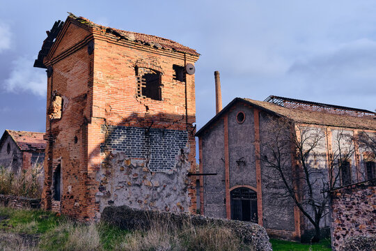 View of a decaying red brick structure with missing roof tiles and broken windows stands against a cloudy sky, contrasting with the green grass, Puertollano, Castile-La Mancha, Spain.