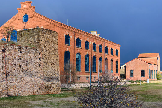 View of the old coal loading facility, a red brick building standing against a moody sky, contrasts with the rustic stone wall, Puertollano, Spain.