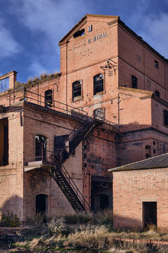 View of a weathered brick building, its windows dark and vacant, stairs ascending against the aged facade under a blue sky, Puertollano, Castile-La Mancha, Spain.