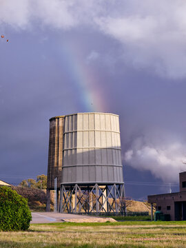 View of a cooling tower standing tall against a stormy sky, a faint rainbow bridging the gap, a stark contrast of industrial and natural beauty, Puertollano, Castile-La Mancha, Spain.