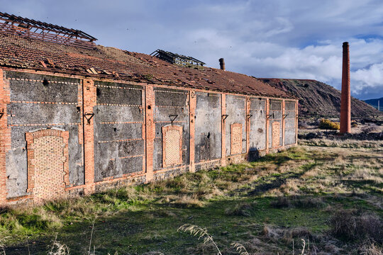 View of the weathered facade of an old coal loading facility stands in stark contrast to the wild grasses and distant smokestack, Puertollano, Castile-La Mancha, Spain.