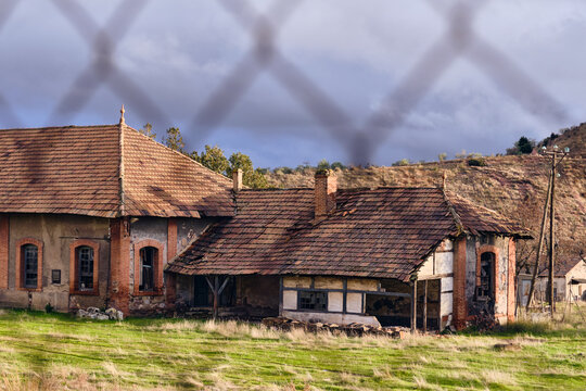 View of abandoned buildings with decaying orange tiled roofs and weathered stone facades rise from a vibrant green field, framed by a chain-link fence, Puertollano, Castile-La Mancha, Spain.