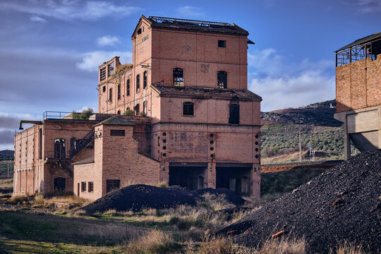 View of the decaying brick facade of an old coal loading facility, with piles of coal, under a cloudy sky, Puertollano, Castile-La Mancha, Spain.