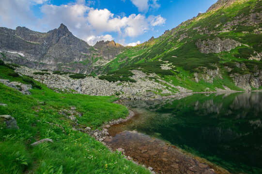 View of jagged peaks rise above a serene lake reflecting verdant slopes and rocky terrain under a bright sky, Zabia Bielovodska dolina, Vysoke Tatry, Presovsky kraj, Slovakia.
