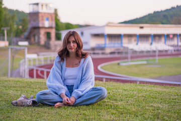 Obraz premium Young woman sitting on the green grass in a city park with a stadium in the background. Peaceful summer evening in the city, lifestyle and relaxation concept. Woman enjoying leisure time outdoors