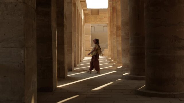 Tourist walks through open-air ancient Egyptian temple with massive columns and shadows cast by sunlight from pillars onto floor