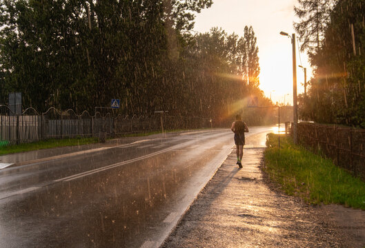 Krakow, Poland &ndash; September 13, 2025: A man runs along a wet roadside during heavy rain at sunset, with sunlight breaking through dark clouds and reflecting on the asphalt after a summer storm.