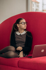 Obraz premium Teen schoolgirl with glasses sitting by red background beside laptop exploring back to deep reading movement attention span crisis print superiority cognitive psychology and education technology shift