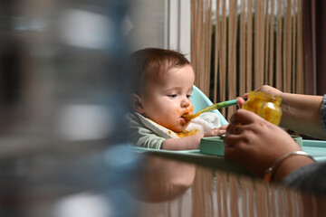 young child in a chair eating clumsily with a spoon, with a blurry reflection on the side