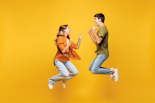 Full body side view young couple friends family man woman wear casual clothes together jump high do winner gesture clench fist isolated on pastel plain light yellow color background studio portrait