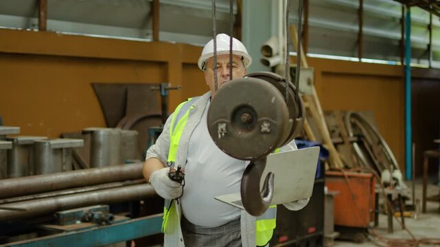 Senior industrial worker operating an overhead crane hook using a remote control while holding a laptop in a factory. Expert technician wearing a safety vest and hard hat in a manufacturing plant.