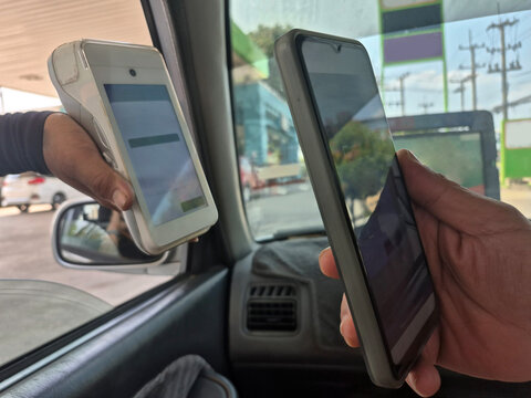 Driver inside a car using a smartphone to scan a QR code on a handheld POS terminal held by an attendant. Concept of contactless digital payment and convenience at a gas station.