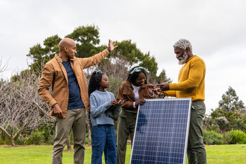 African American family standing on grassy lawn, examining freestanding solar panel, holding cables