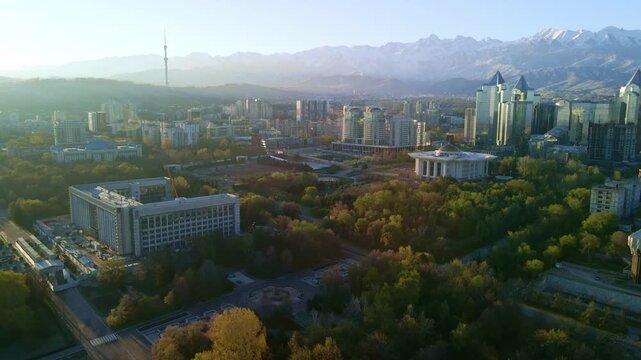 Cinematic aerial drone flight over Almaty city skyline. Panoramic view of modern skyscrapers and historical buildings with snow-capped Tian Shan mountains in Kazakhstan.