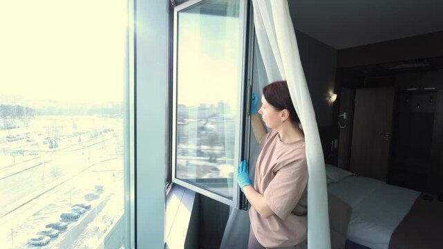 A maid in a hotel room, wearing gloves, opens the window to let in fresh air.
