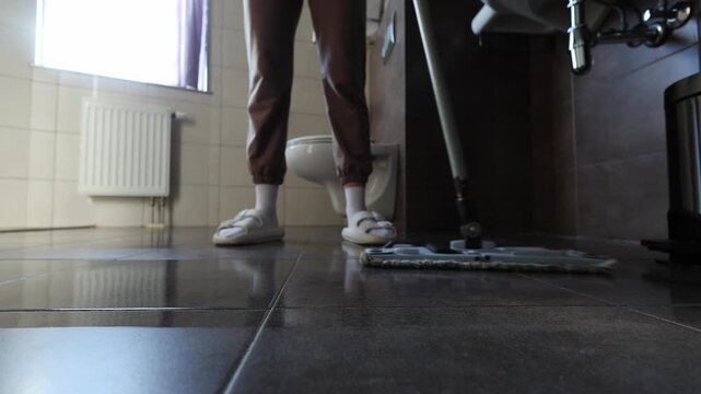 A person mops a tiled bathroom floor, focusing on the area near the toilet and sink.