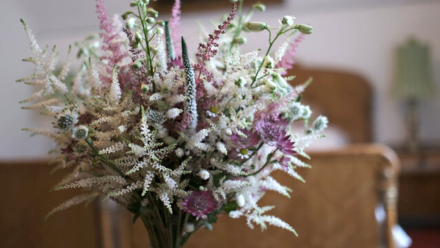 A delicate and airy floral arrangement featuring soft pink and white wildflowers, astilbe, and masterwort in a blurrily lit indoor setting.