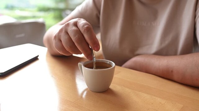 Close-up video of a person stirring coffee with a spoon in a white cup on a wooden table. Cozy indoor scene with soft natural light.
