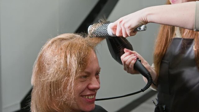 Stylist forms perfect curls at ends of short wet hair of beautiful woman after keratin treatment in salon. Towel removed, she smiles confidently in mirror, premium beauty and self-care experience.