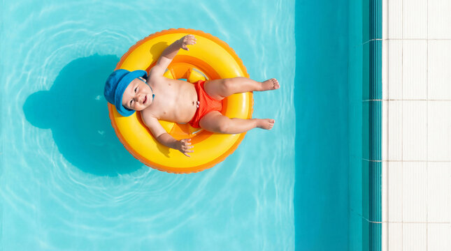 Happy baby smiling and relaxing in a bright yellow infant swim ring floating on clean turquoise pool water top down view, for summer vacation ads, poolside baby happiness content