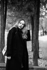 Female model in black coat and knee-high boots stands among autumn leaves on a tree-lined path, showcasing a stylish look in a monochrome setting