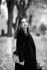Female model in black coat and knee-high boots stands among autumn leaves on a tree-lined path, showcasing a stylish look in a monochrome setting