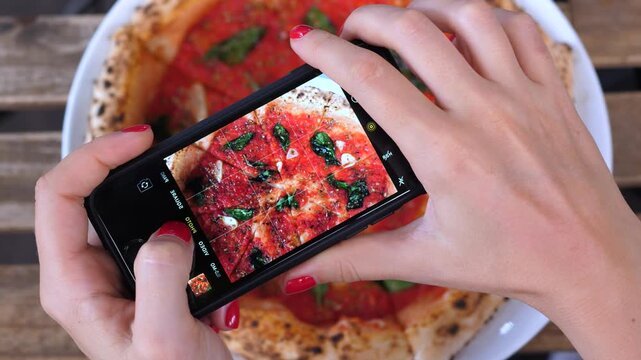 Female hands holding a smartphone, taking a picture of a fresh italian pizza for social media. Food blogger creating content in a restaurant, capturing a delicious meal from a top down view
