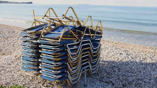 Stack of sunbeds chaise-longues piled up on the pebble beach, preparing beach for opening season,  the end of touristic season in Greece, pile of stacked deck chairs, Ionian sea islands, high season i