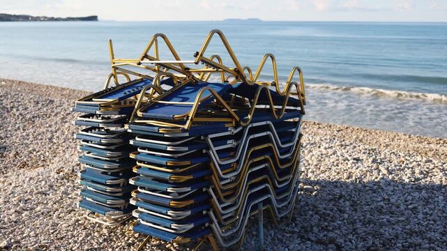 Stack of sunbeds chaise-longues piled up on the pebble beach, preparing beach for opening season,  the end of touristic season in Greece, pile of stacked deck chairs, Ionian sea islands, high season i