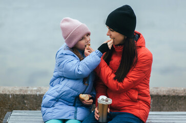 Photograph, portrait of a teenage girl with her mother, feeding each other a croissant and hot tea in the cold while sitting on a bench on the embankment with the sea in the background.