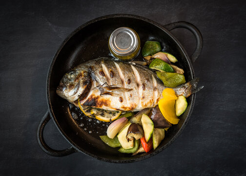 A cooked sea bream fish in a pan with mixed vegetables on a dark background in a restaurant
