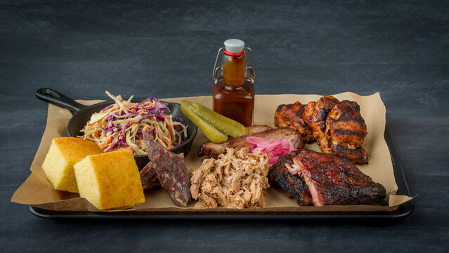 A classic barbecue meat platter containing spare ribs, pulled pork, smoked spicy sausage, beef brisket slices, grilled chicken wings and drumsticks, coleslaw, corn bread, pickles and bbq sauce