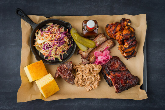 Top view of a classic barbecue meat platter containing spare ribs, pulled pork, smoked spicy sausage, beef brisket slices, grilled chicken wings and drumsticks, coleslaw, corn bread, pickles and sauce