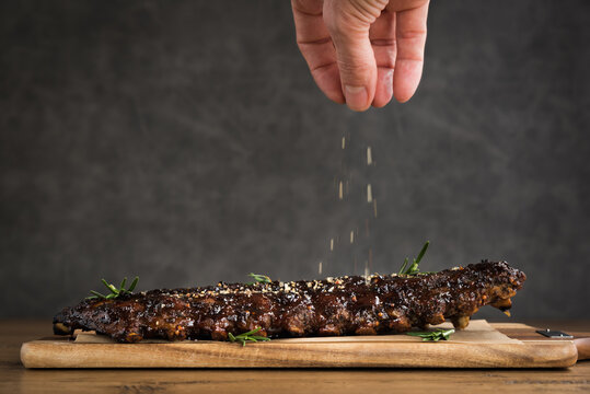A rack of barbecued spare ribs covered in sauce on a wooden plank with a dark background and a hand dropping extra herbs and seasoning on top of ribs