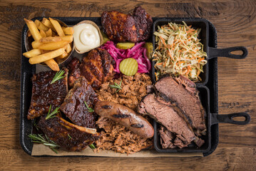 Top view of a barbecue meat platter with ribs, pulled pork, sausage, beef brisket slices, coleslaw, fires, mayonnaise, chicken pieces and pickles