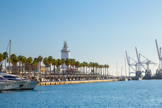 Pier One Muelle Uno waterfront promenade with palm trees, La Farola lighthouse, and industrial port cranes in the background under a clear Mediterranean sky with urban coastal landscape.