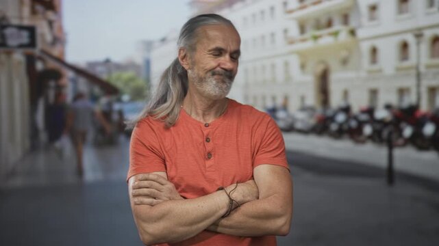 Man with folded arms and visible forearms standing on european street, long hoary hair and beard; confidence maturity wisdom.