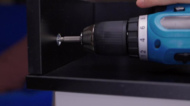 Close-up of person driving screw with cordless drill into black furniture panel, assembling drawer on workbench with tools and hardware in dark workshop interior, close-up, slow motion.