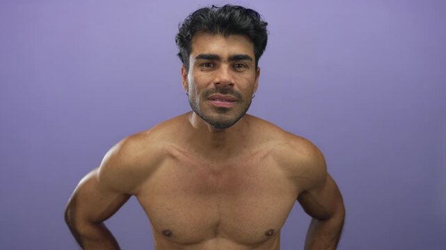 Young hispanic man leaning forward with bare chest and hands on hips in purple studio; physical discomfort.