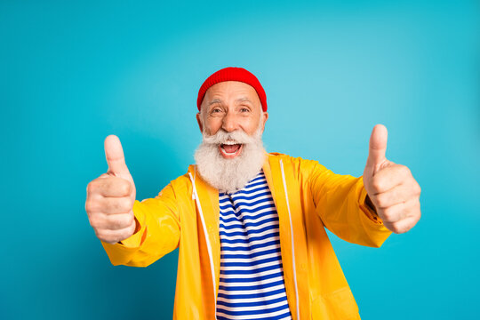 Happy bearded senior man in yellow raincoat gives thumbs up against blue background