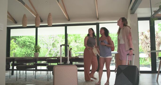Diverse female friends arriving at vacation home, pulling suitcases, exploring layout, hugging